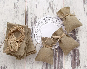 May include: An overhead shot of several burlap items on a white, distressed wooden surface. There are small burlap bags tied with twine, stacks of burlap squares, and a bundle of twine. A white paper doily is also present.
