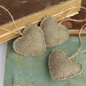 Puede incluir: Tres corazones de arpillera marr&oacute;n con una cuerda de yute r&uacute;stica atada a cada uno. Los corazones est&aacute;n dispuestos sobre una pila de libros antiguos.