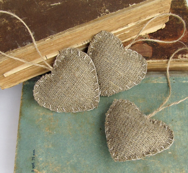 Puede incluir: Tres corazones de arpillera marr&oacute;n con una cuerda de yute r&uacute;stica atada a cada uno. Los corazones est&aacute;n dispuestos sobre una pila de libros antiguos.
