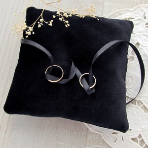 May include: A square black velvet ring pillow featuring a central tufted detail and two thin black satin ribbons tied to hold two plain gold-toned metal bands. A small sprig of dried white baby's breath flowers rests on the top edge of the pillow.