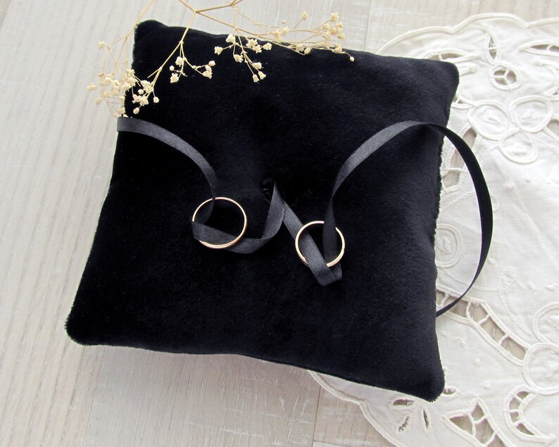 May include: A square black velvet ring pillow featuring a central tufted detail and two thin black satin ribbons tied to hold two plain gold-toned metal bands. A small sprig of dried white baby's breath flowers rests on the top edge of the pillow.