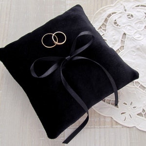 May include: A square black velvet ring pillow featuring a centered black satin ribbon tied into a bow. Two plain gold-toned metal bands are positioned on the top surface of the cushion, resting near the ribbon bow on the soft fabric texture.