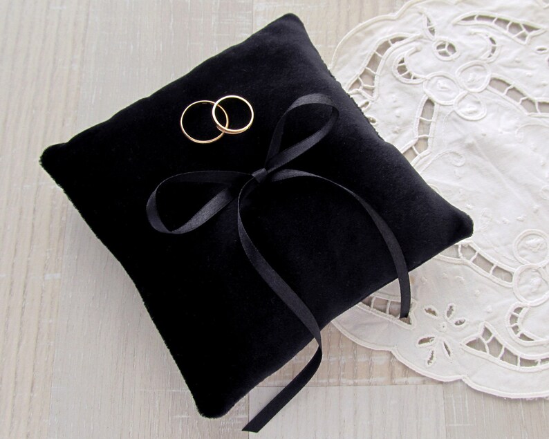 May include: A square black velvet ring pillow featuring a centered black satin ribbon tied into a bow. Two plain gold-toned metal bands are positioned on the top surface of the cushion, resting near the ribbon bow on the soft fabric texture.