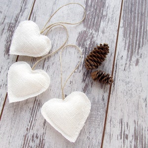 Puede incluir: Tres adornos de coraz&oacute;n de tela blanca con colgadores de cuerda en un fondo de madera blanqueada. Tambi&eacute;n hay dos pi&ntilde;as en la superficie de madera.