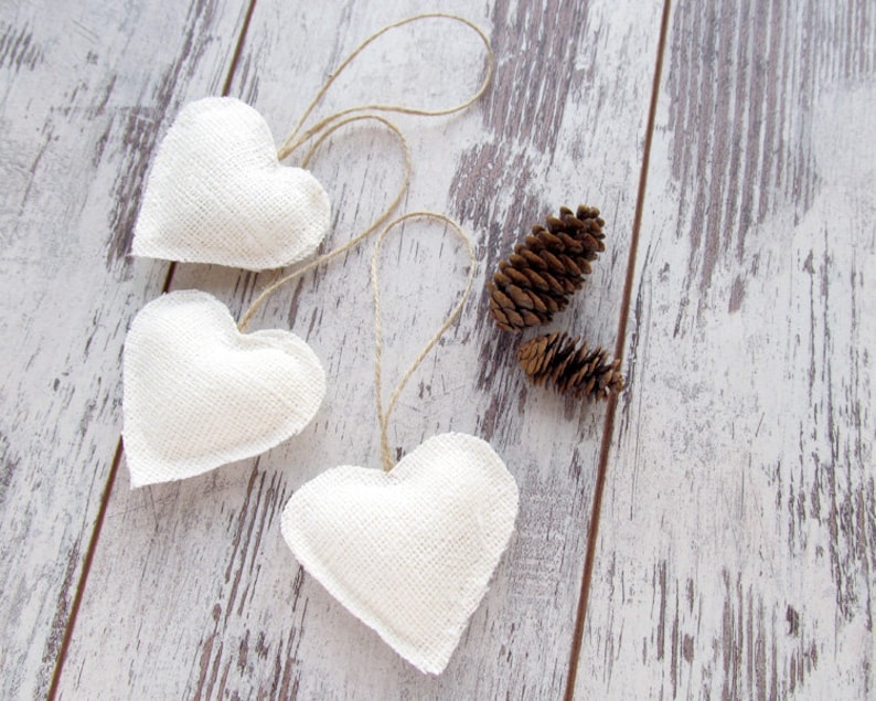 Puede incluir: Tres adornos de coraz&oacute;n de tela blanca con colgadores de cuerda en un fondo de madera blanqueada. Tambi&eacute;n hay dos pi&ntilde;as en la superficie de madera.
