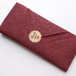 May include: A rectangular envelope made of deep burgundy textured paper. The envelope is sealed with a round, scalloped-edge tan sticker featuring the black text Thank you! and a small red heart icon centered on the flap of the envelope.