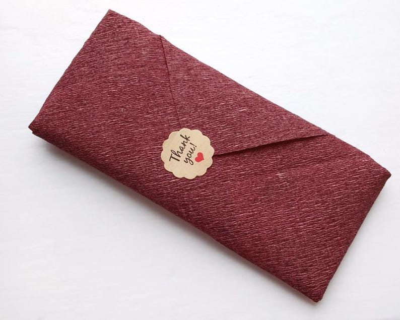 May include: A rectangular envelope made of deep burgundy textured paper. The envelope is sealed with a round, scalloped-edge tan sticker featuring the black text Thank you! and a small red heart icon centered on the flap of the envelope.