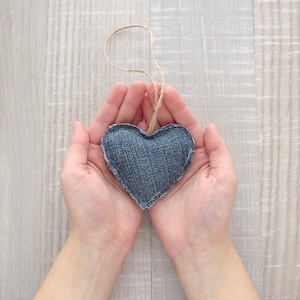 Puede incluir: Dos manos sosteniendo una almohada de coraz&oacute;n de mezclilla azul con una cuerda atada.