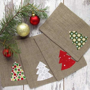 May include: Four square, tan linen napkins with appliqued Christmas tree designs in red, white, and green patterns. A sprig of pine with red and gold ornaments is in the background.