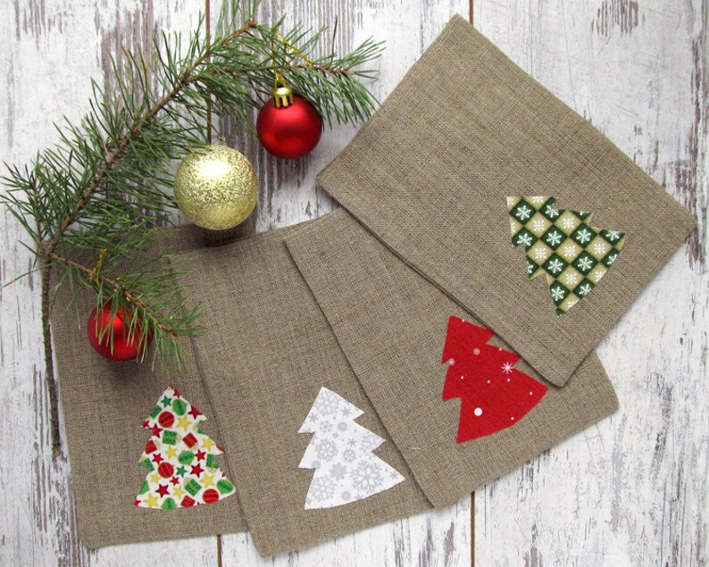 May include: Four square, tan linen napkins with appliqued Christmas tree designs in red, white, and green patterns. A sprig of pine with red and gold ornaments is in the background.