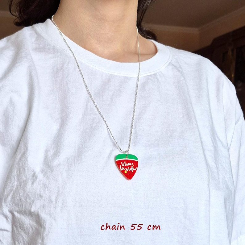 May include: A silver chain necklace with a pendant shaped like a strawberry. The pendant is red with a green top and the words "Viva la vida" in white script. The chain is 21.65 inches long.