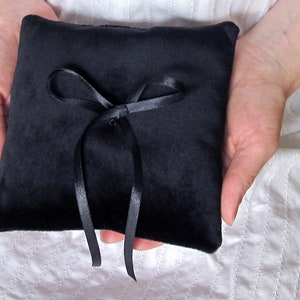 May include: A square ring bearer pillow made of black velvet fabric. The center of the pillow features a decorative black satin ribbon tied into a bow, with two long ribbon tails extending downward from the knot, providing a secure attachment point.