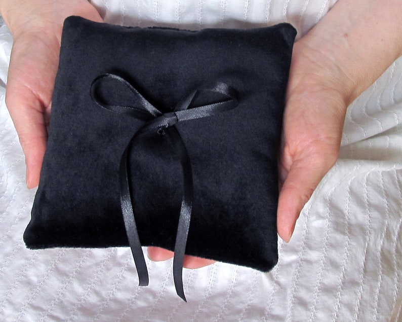 May include: A square ring bearer pillow made of black velvet fabric. The center of the pillow features a decorative black satin ribbon tied into a bow, with two long ribbon tails extending downward from the knot, providing a secure attachment point.
