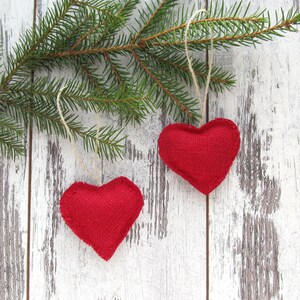 Puede incluir: Dos corazones de tela roja colgando de una rama de pino sobre un fondo de madera blanqueada.