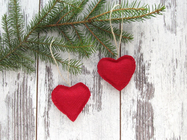 Puede incluir: Dos corazones de tela roja colgando de una rama de pino sobre un fondo de madera blanqueada.