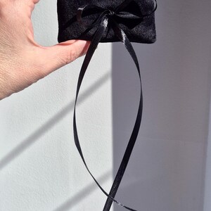 May include: A small, rectangular black velvet pillow featuring a centered black satin ribbon tied into a bow. The ribbon has two long, dangling ends that extend downward from the bow, creating a simple and elegant presentation for small items.