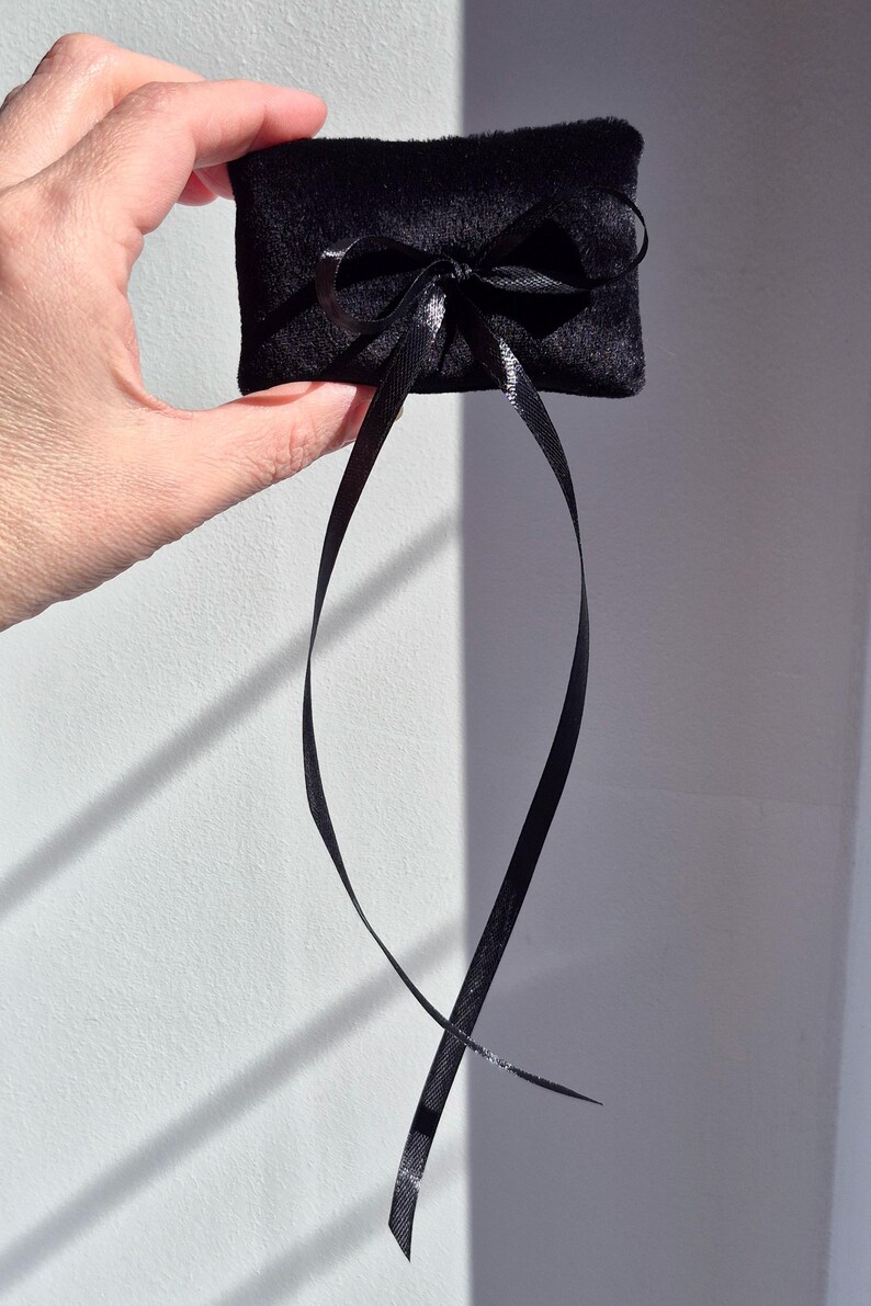 May include: A small, rectangular black velvet pillow featuring a centered black satin ribbon tied into a bow. The ribbon has two long, dangling ends that extend downward from the bow, creating a simple and elegant presentation for small items.
