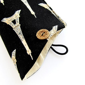 May include: A black fabric pouch featuring a repeating white and gold Eiffel Tower pattern. The pouch has a light beige interior lining, a circular wooden button closure with a floral design, and a black elastic loop for securing the flap.