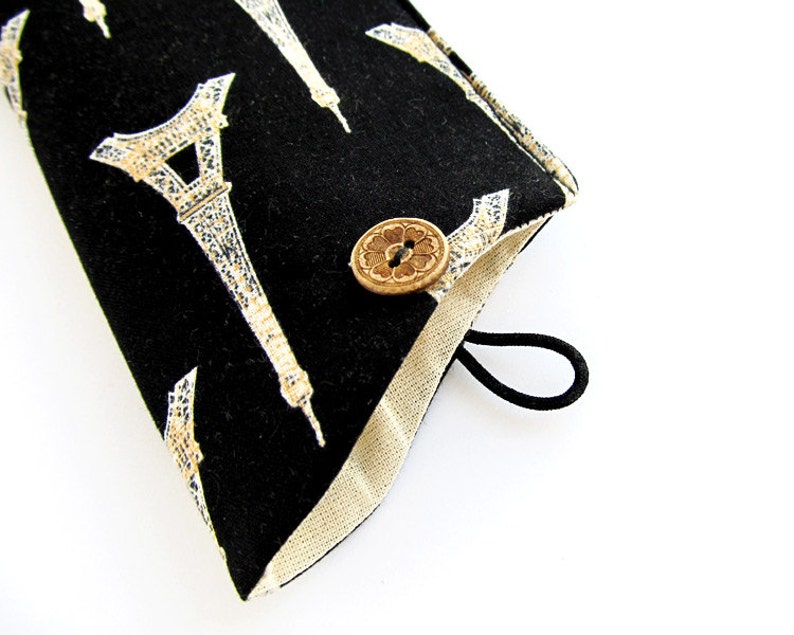 May include: A black fabric pouch featuring a repeating white and gold Eiffel Tower pattern. The pouch has a light beige interior lining, a circular wooden button closure with a floral design, and a black elastic loop for securing the flap.