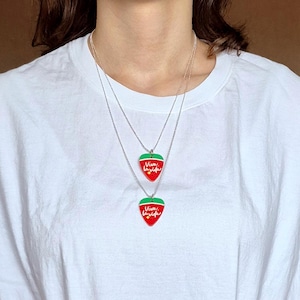 May include: Two silver necklaces with red strawberry pendants. Each pendant has a green top and the words "Viva la vida" in red script. The necklaces are layered and worn against a white t-shirt.