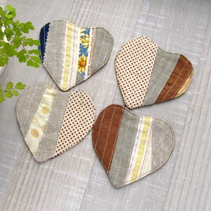 Puede incluir: Cuatro posavasos de tela en forma de corazón con diseños de patchwork. Cada posavasos presenta una combinación única de patrones, incluyendo lunares, rayas y estampados florales. La paleta de colores incluye beige, marrón, amarillo y azul.