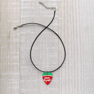 May include: A black choker necklace with a guitar pick pendant. The pendant is red with a green top and features the text "Viva la vida" in white script. The necklace has a silver-colored clasp and chain.
