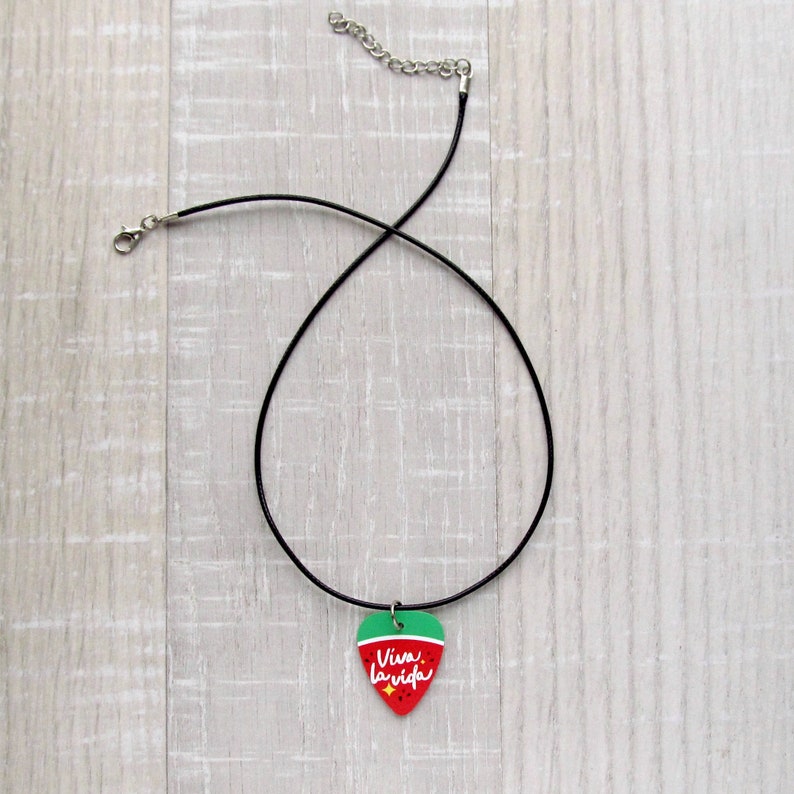 May include: A black choker necklace with a guitar pick pendant. The pendant is red with a green top and features the text "Viva la vida" in white script. The necklace has a silver-colored clasp and chain.