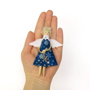 Op de afbeelding: Een blauwe en witte stoffen engelversiering met een sneeuwvlokkenbedel. De engel heeft blond haar en witte vleugels.
