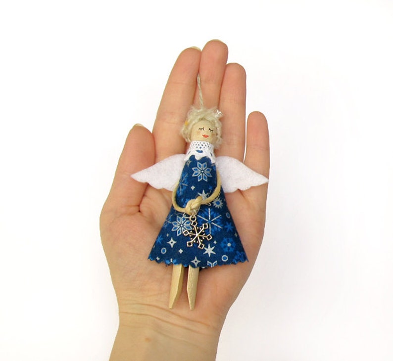 Op de afbeelding: Een blauwe en witte stoffen engelversiering met een sneeuwvlokkenbedel. De engel heeft blond haar en witte vleugels.