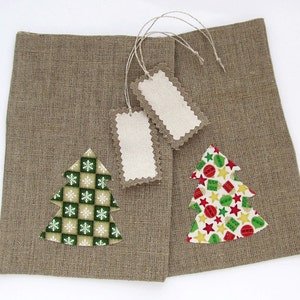 May include: Two brown burlap gift bags with Christmas tree appliqu&eacute;s. One tree has a green and white snowflake pattern, the other has a colorful star and ornament pattern. Two blank tags with scalloped edges are attached.