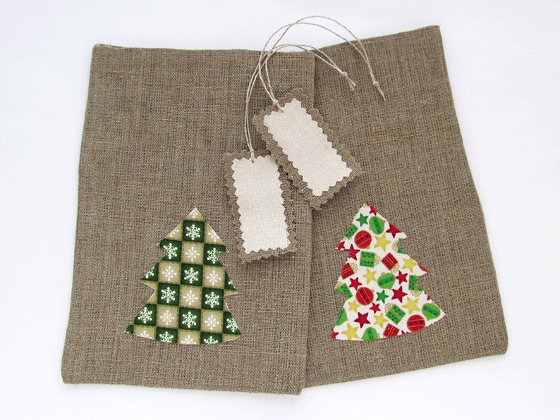 May include: Two brown burlap gift bags with Christmas tree appliqu&eacute;s. One tree has a green and white snowflake pattern, the other has a colorful star and ornament pattern. Two blank tags with scalloped edges are attached.