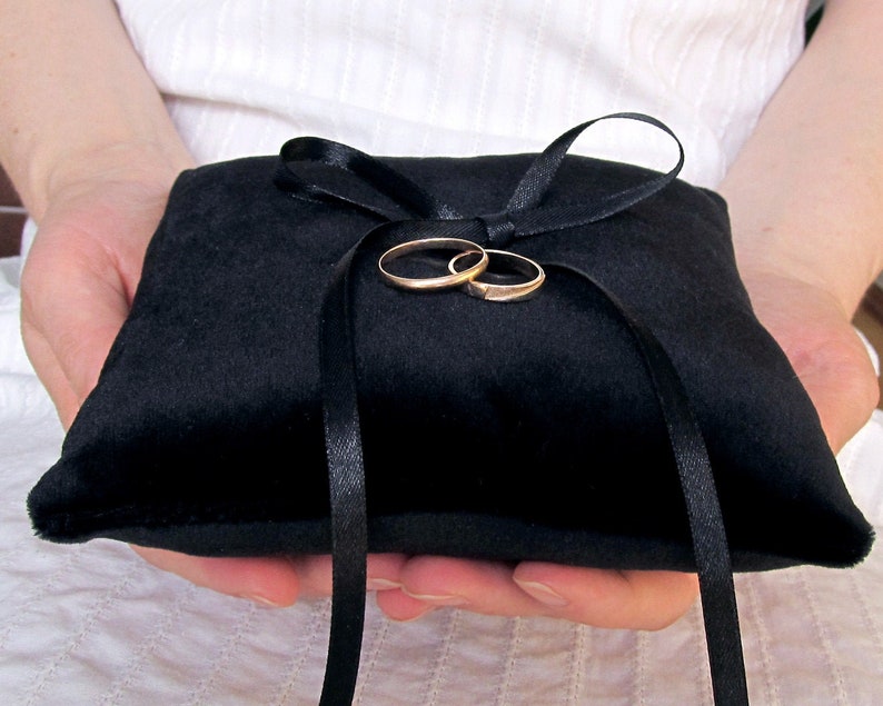 May include: A square black velvet ring pillow featuring a smooth texture and a centered black satin ribbon tied into a bow. Two plain gold-toned metal bands are resting securely on the top of the pillow, held in place by the ribbon loops.