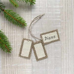 May include: Three rectangular gift tags with a natural linen center and a decorative brown border. One tag has the name "Diane" written in black script. The tags are attached to twine and are next to a green pine branch.