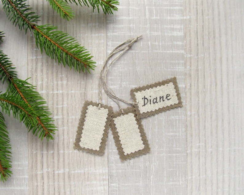 May include: Three rectangular gift tags with a natural linen center and a decorative brown border. One tag has the name "Diane" written in black script. The tags are attached to twine and are next to a green pine branch.