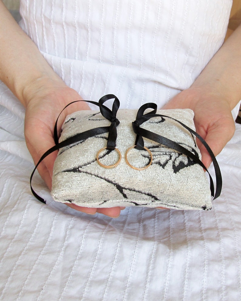 May include: A square ring pillow featuring a textured cream and black leaf-patterned fabric. Two thin black satin ribbons are tied in bows on the top surface, each holding a simple gold-toned metal ring suspended from the center of the cushion.