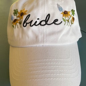 May include: White baseball cap with embroidered flowers and the word "bride" in black cursive.