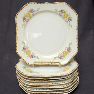 May include: A stack of six white square plates with scalloped edges and a gold trim. Each plate features a floral design with yellow, pink, and blue flowers. The plates are stacked on a gold-colored plate stand.