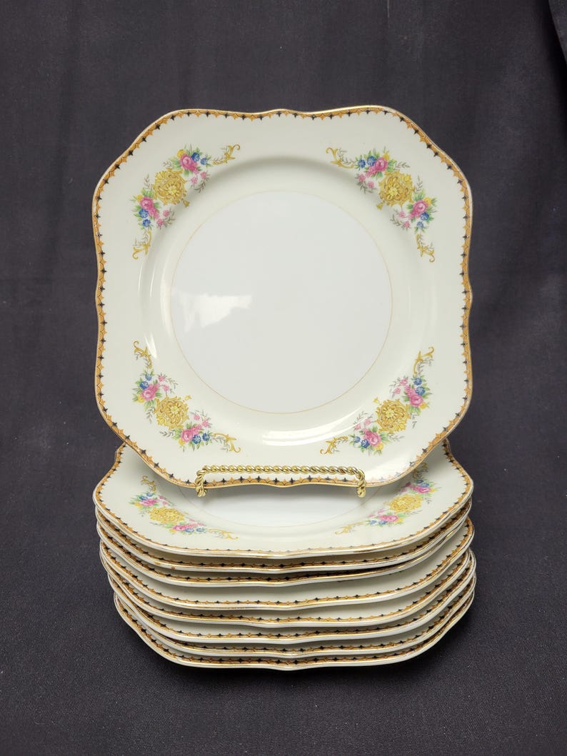 May include: A stack of six white square plates with scalloped edges and a gold trim. Each plate features a floral design with yellow, pink, and blue flowers. The plates are stacked on a gold-colored plate stand.