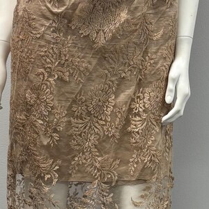 Pode incluir: Um vestido longo e fluido com um delicado padr&atilde;o de renda floral em cor castanho claro. O vestido &eacute; sem mangas e tem um corpete justo com uma sobreposi&ccedil;&atilde;o de renda em camadas.