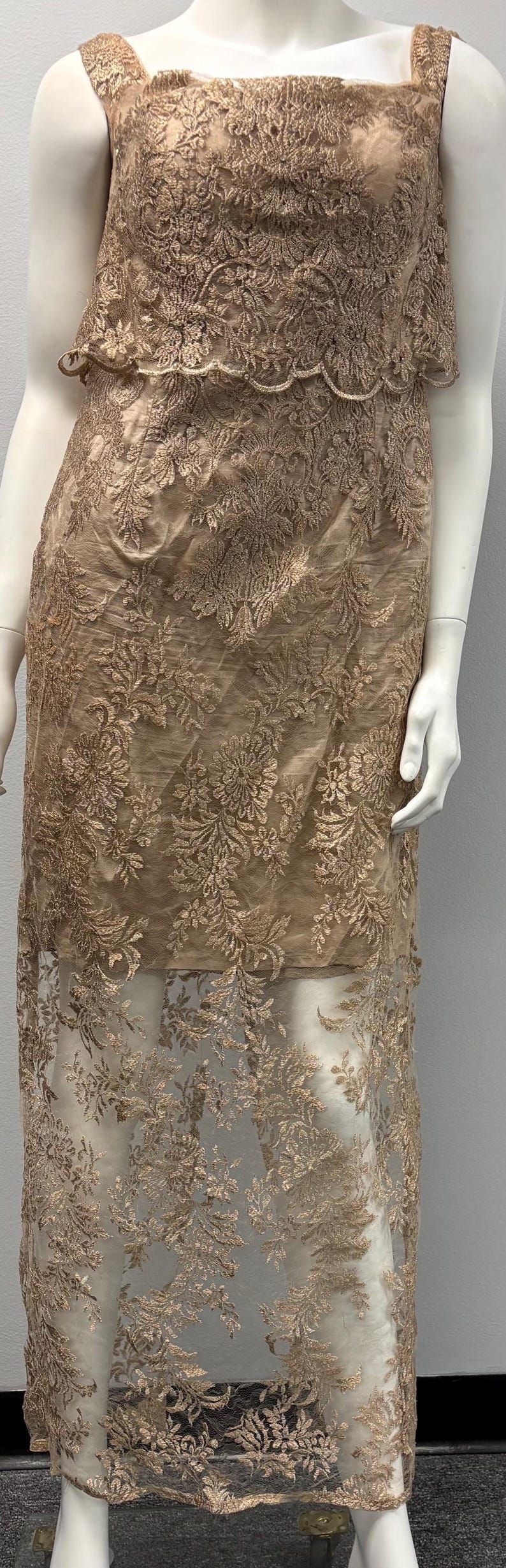 Pode incluir: Um vestido longo e fluido com um delicado padr&atilde;o de renda floral em cor castanho claro. O vestido &eacute; sem mangas e tem um corpete justo com uma sobreposi&ccedil;&atilde;o de renda em camadas.