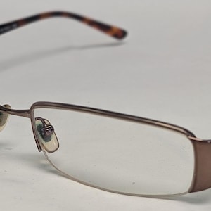 May include: A pair of eyeglasses with a brown metal frame and clear lenses. The temples are a tortoiseshell pattern, and the frame has a rectangular shape. The brand name is visible on the temple.