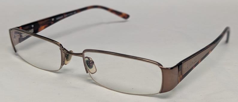May include: A pair of eyeglasses with a brown metal frame and clear lenses. The temples are a tortoiseshell pattern, and the frame has a rectangular shape. The brand name is visible on the temple.