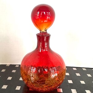 May include: A red glass decanter with a round body and a spherical stopper. The decanter has a crackle finish on the lower half, transitioning to a smooth, glossy red on top. The stopper is a vibrant red-orange sphere.