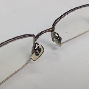 May include: Close-up of eyeglasses with a thin, dark metal frame. The lenses are clear, and the nose pads are visible. The glasses are set against a white background, highlighting the details of the eyewear.
