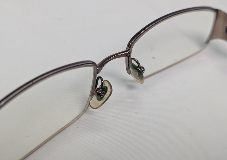 May include: Close-up of eyeglasses with a thin, dark metal frame. The lenses are clear, and the nose pads are visible. The glasses are set against a white background, highlighting the details of the eyewear.