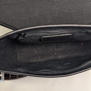 May include: Open view of a black leather crossbody bag. The interior is black with a zippered pocket and a small leather tag. The bag has a black fabric flap and a silver buckle.