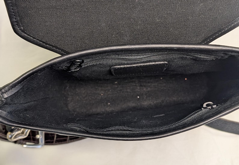 May include: Open view of a black leather crossbody bag. The interior is black with a zippered pocket and a small leather tag. The bag has a black fabric flap and a silver buckle.