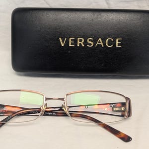 May include: Eyeglasses with a tortoiseshell pattern and a gold-colored frame, resting in front of a black leather case. The case has the brand name "VERSACE" in gold lettering.