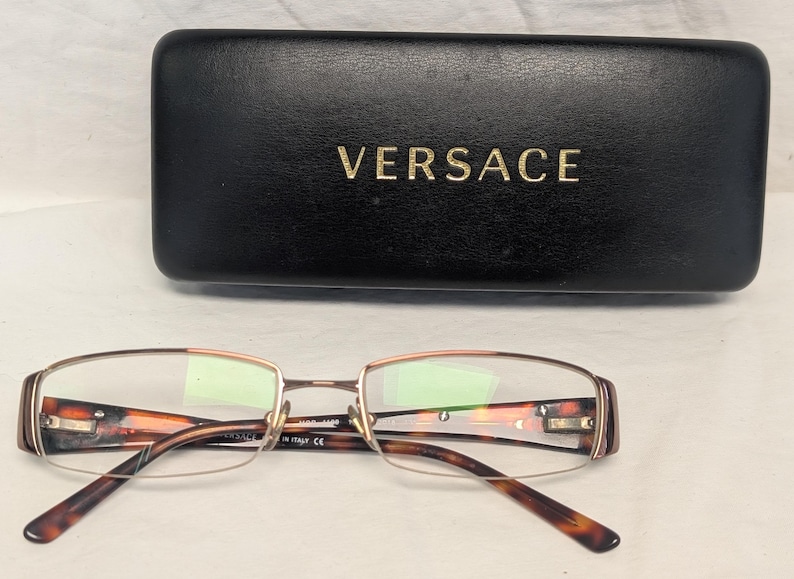 May include: Eyeglasses with a tortoiseshell pattern and a gold-colored frame, resting in front of a black leather case. The case has the brand name "VERSACE" in gold lettering.