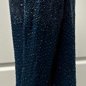 May include: A long-sleeved, dark teal beaded top. The garment is covered in small, shimmering beads that create a textured effect. The beads are densely packed at the top and gradually become less dense towards the bottom.